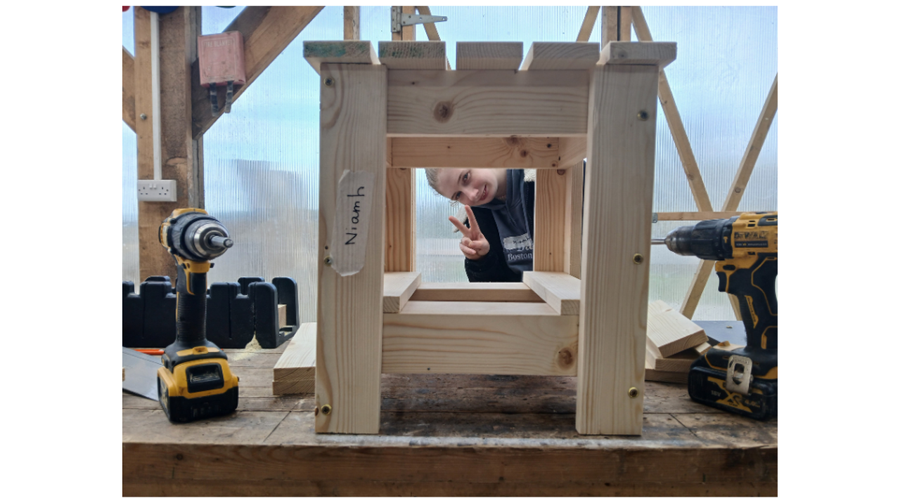 A wooden stool under construction on a workbench surrounded by drills, with a person playfully peeking through and making a peace sign.